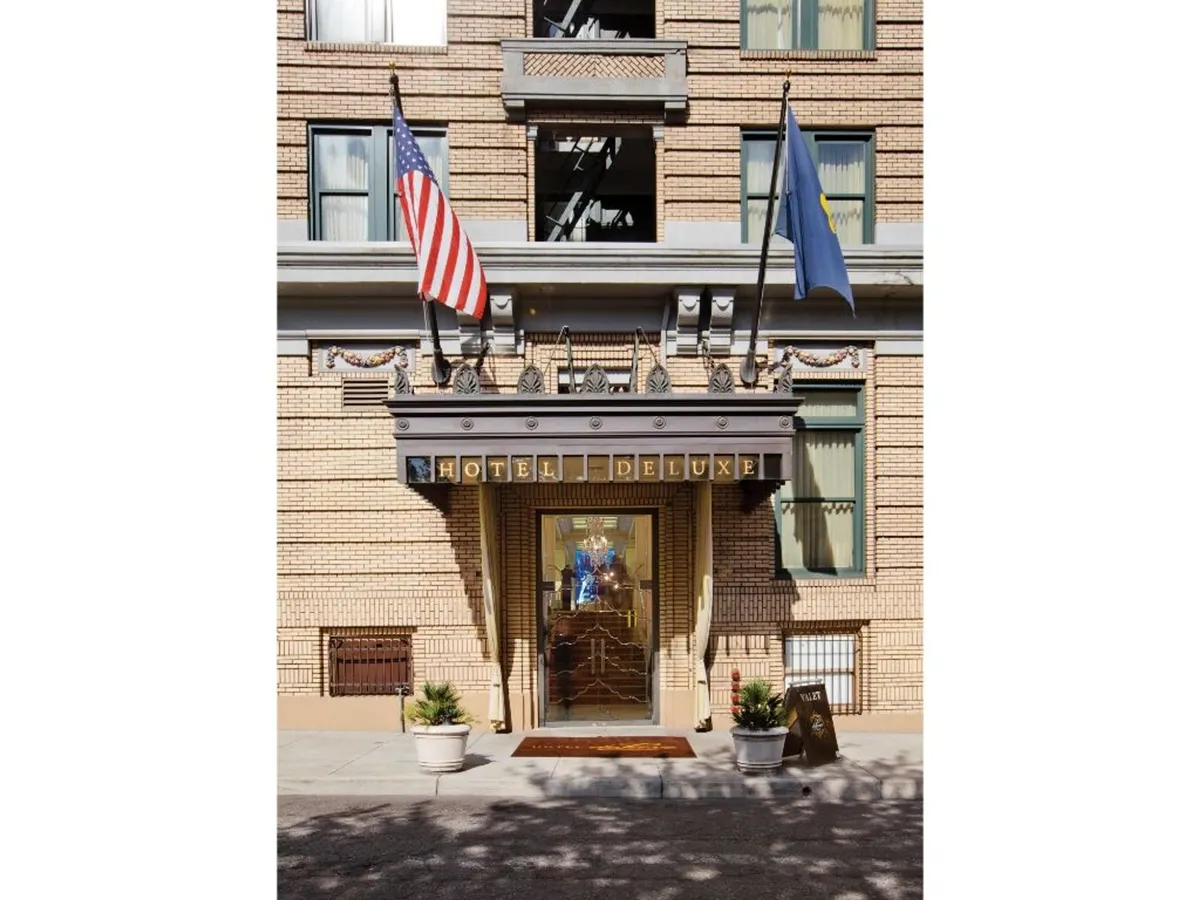 This image shows the entrance to Hotel Deluxe, featuring a clear view of the doorway, hotel sign, and multiple flags. The scene captures guests arriving and socializing, which highlights the accessible entry point. The setting looks welcoming and visually establishes the hotel's location as a gateway to exploring downtown Portland.