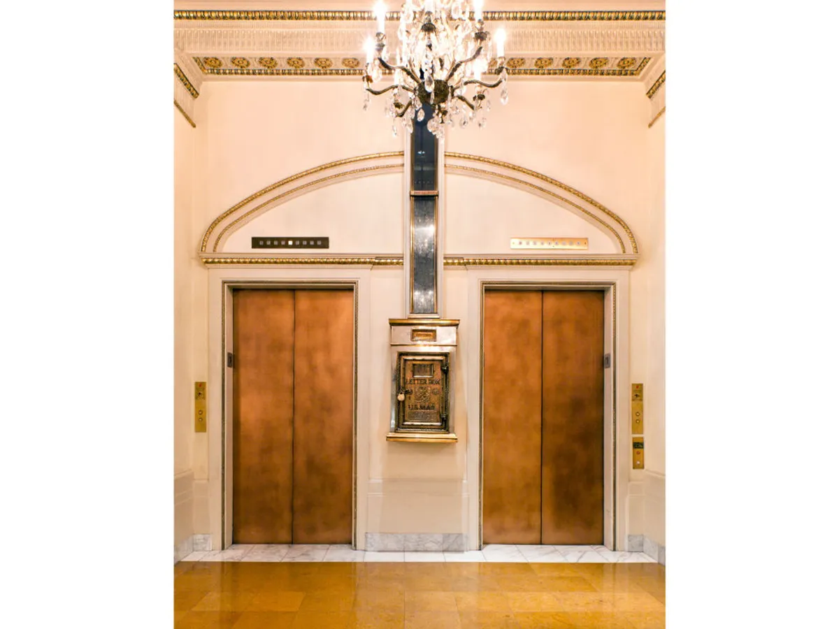 A distinguished lobby scene with ornate chandelier, decorative wall moldings, and elevator doors, capturing the sense of arrival and anticipation. The mood is both welcoming and grand, reflecting the first impression of the property.