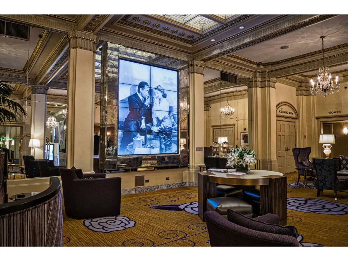 An elegant lounge adorned with a decorative carpet, a chandelier, comfortable seating, and a large display screen featuring a black-and-white photo. The space is designed for conversation and unwinding in vintage-inspired luxury.