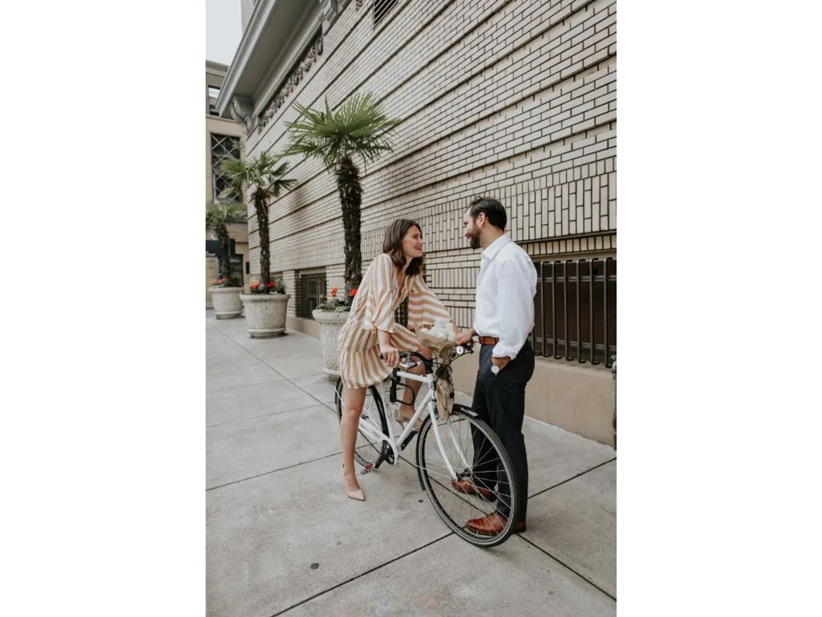 An exterior scene of a couple interacting by a bicycle outside a brick wall and potted plants, expressing joy and spontaneity. The image highlights the experience of borrowing a complimentary bike and exploring downtown Portland.