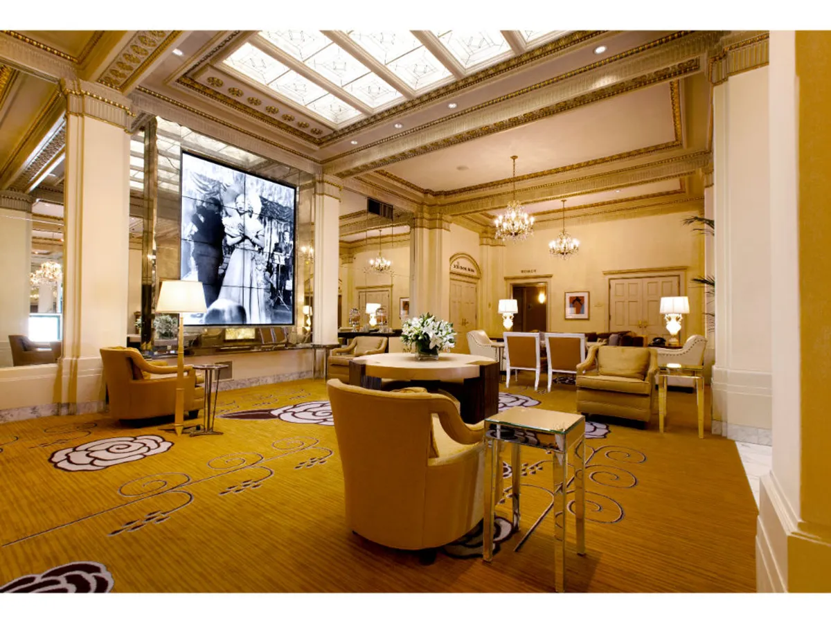Inside the hotel lobby, stylish seating areas, ornate chandeliers, decorative carpets, and a large digital screen displaying a historical photograph set a sophisticated tone. The space is both welcoming for relaxation and ideal for socializing or art appreciation.