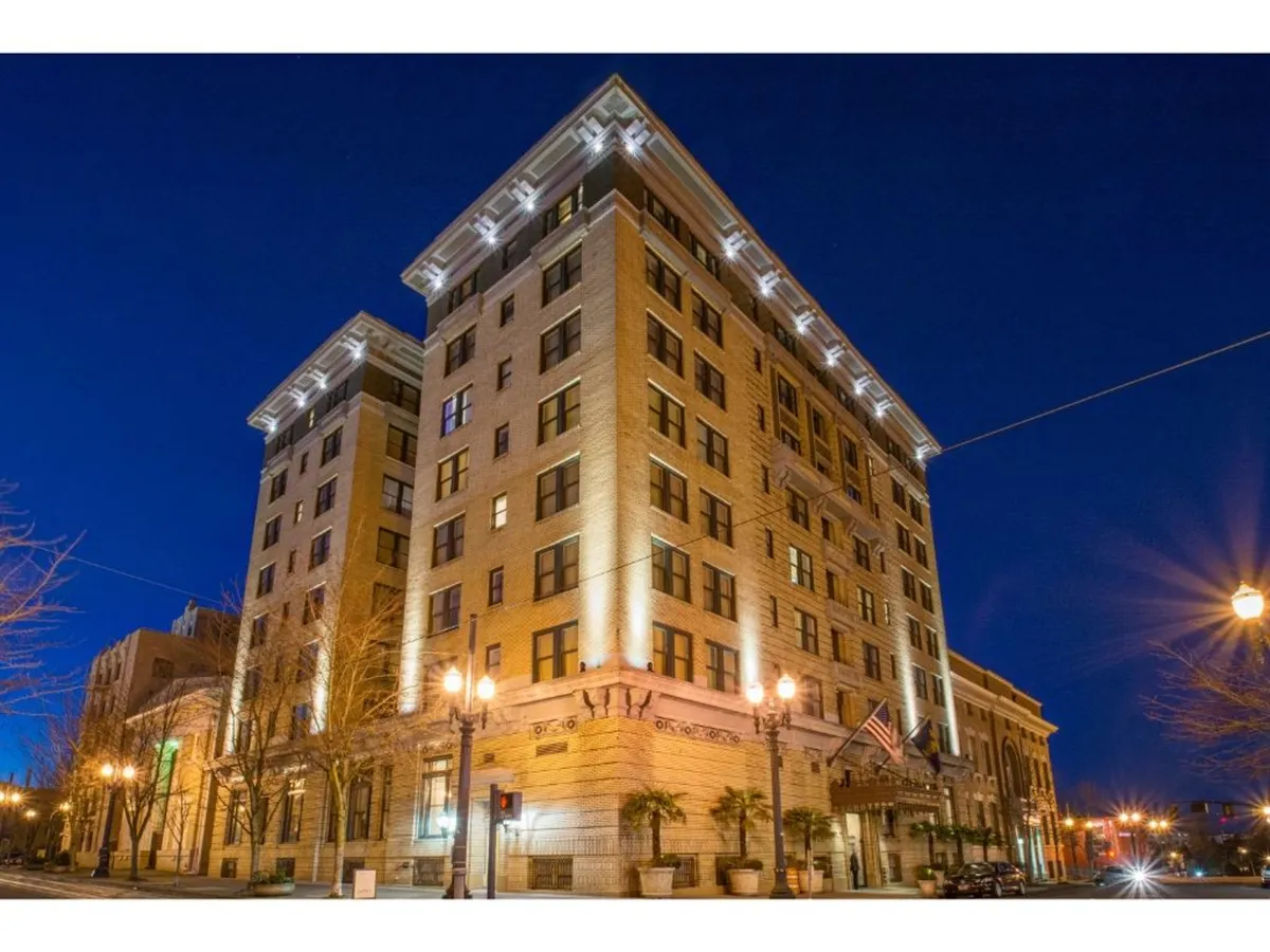 An evening exterior view of the hotel's historic architecture, with illuminated columns, palm trees, and street lamps. The scene is alive with sophisticated energy, suggesting easy access to city nightlife, dining, and iconic attractions right outside the door.