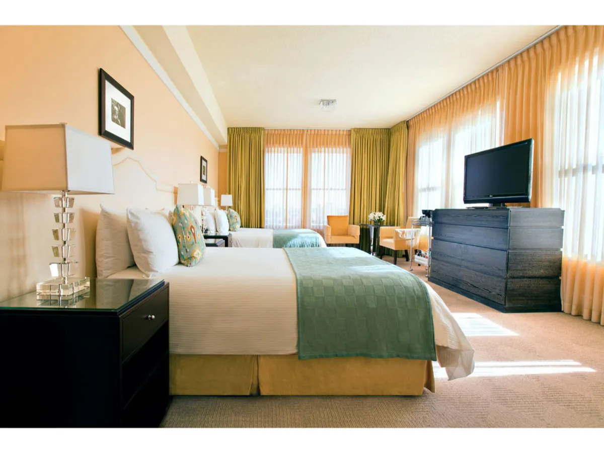 This image showcases a serene guest room featuring two queen beds with colorful throws, a sitting area with a chair and table, large windows with sheer curtains allowing sunlight to stream in, and a modern television unit. The atmosphere is welcoming and tranquil, highlighting comfort and thoughtful arrangement.