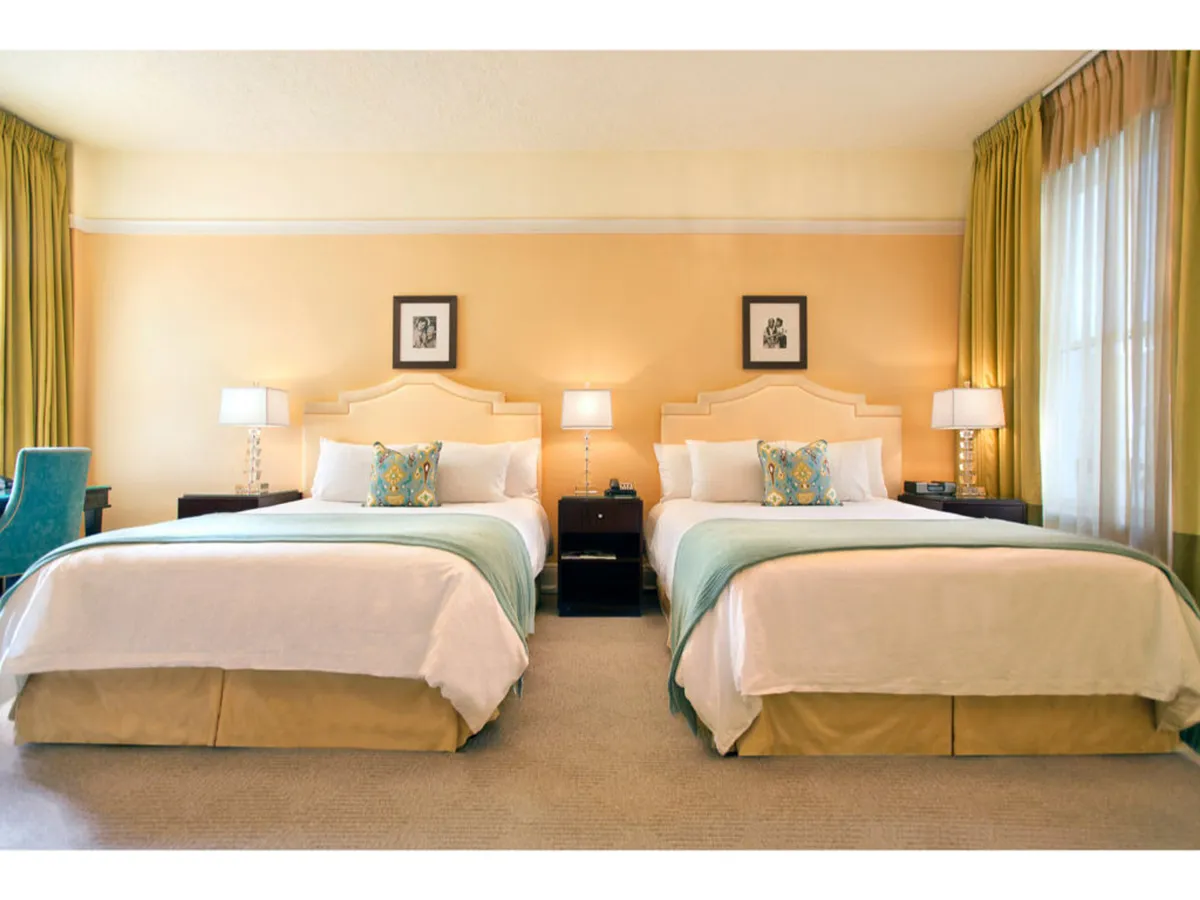 The image showcases a stylish hotel room with two plush double beds adorned with decorative pillows, flanked by elegant nightstands and soft-glowing lamps. Natural light filters through the large window with flowing curtains, creating a bright yet cozy ambiance ideal for relaxation or planning your Portland adventures.