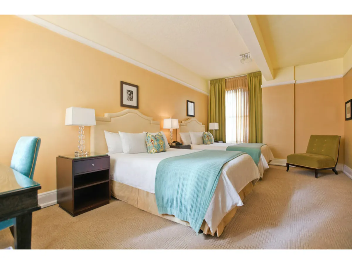 A cozy interior with two beds adorned with decorative pillows, a green accent chair, and a table lamp with a crystal base. Sunlight fills the room, enhancing the welcoming and restful environment.