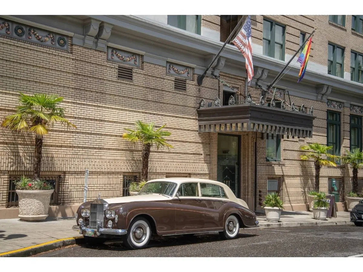 The historic hotel facade is paired with a classic car out front, palm trees, and visible flags. The exterior shot is lively, showing guests arriving and the vibrant street scene, hinting at the hotel's proximity to downtown attractions and accessible city life.