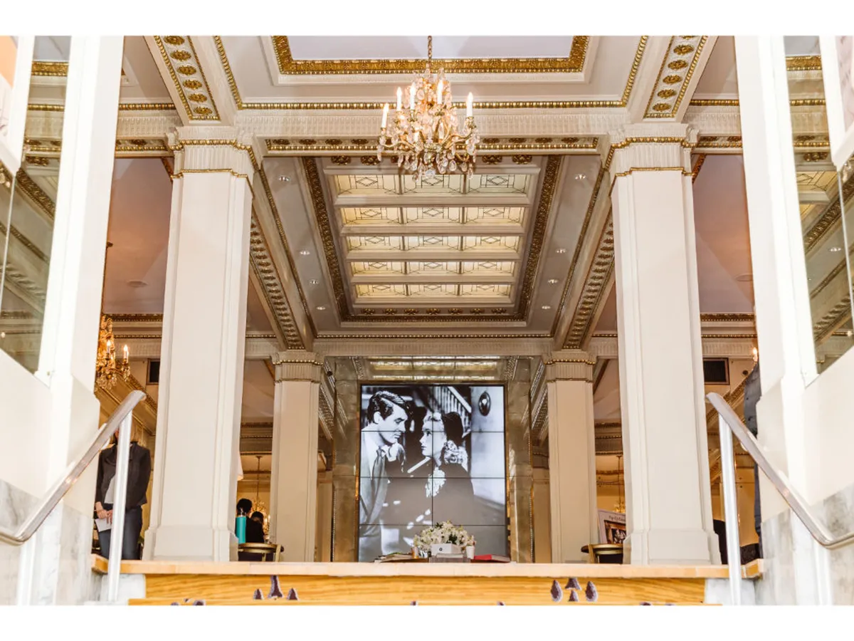 An opulent, inviting interior showcasing a dazzling chandelier, a decorative ceiling, and an impressive black-and-white photo display near a grand staircase. The space is ideal for formal gatherings or art events, blending grandeur with creative energy.