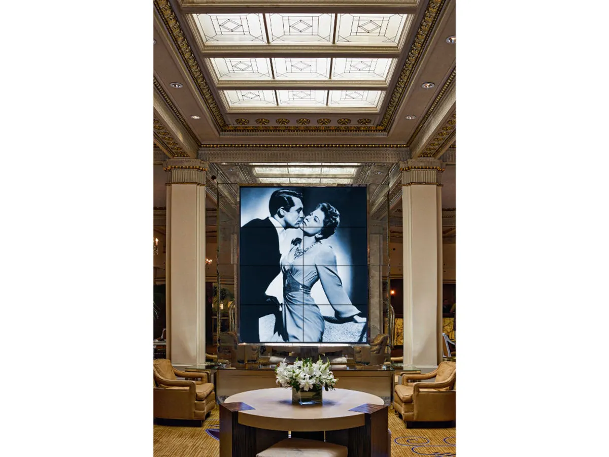 A sophisticated hotel lounge area featuring a striking black and white photograph of a couple, a decorative ceiling with glass panels, and a variety of stylish seating arrangements. The space is designed for intimate gatherings and offers an inviting atmosphere suitable for informal business discussions or unwinding after meetings.